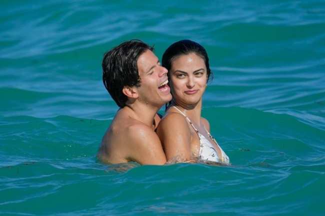 Rudy Mancuso riendo mientras abraza a Camila Mendes por detras en un oceano