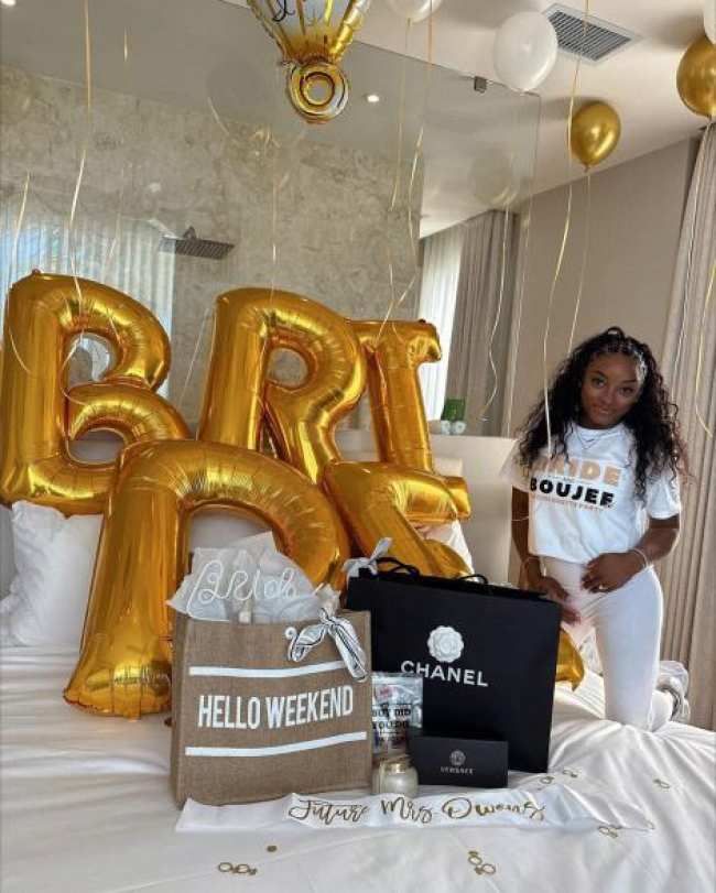 Simone Biles de pie junto a una cama cubierta de globos y regalos de novia