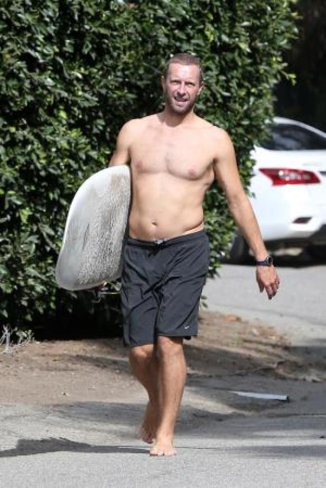 Chris Martin sin camisa que lleva una tabla de surf en pantalones cortos