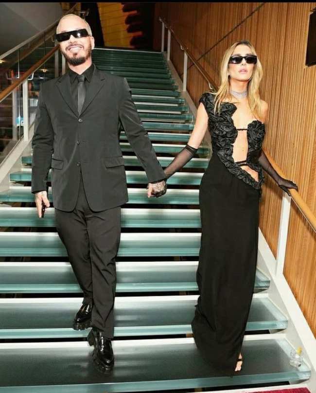 La superestrella latina J Balvin vestido con un traje oscuro y su novia Valentina Ferrer con un vestido negro escotado bajando las escaleras