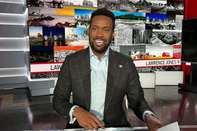 El presentador de Fox News Lawrence Jones vestido con una camisa blanca y una chaqueta gris sonrie detras del escritorio del presentador