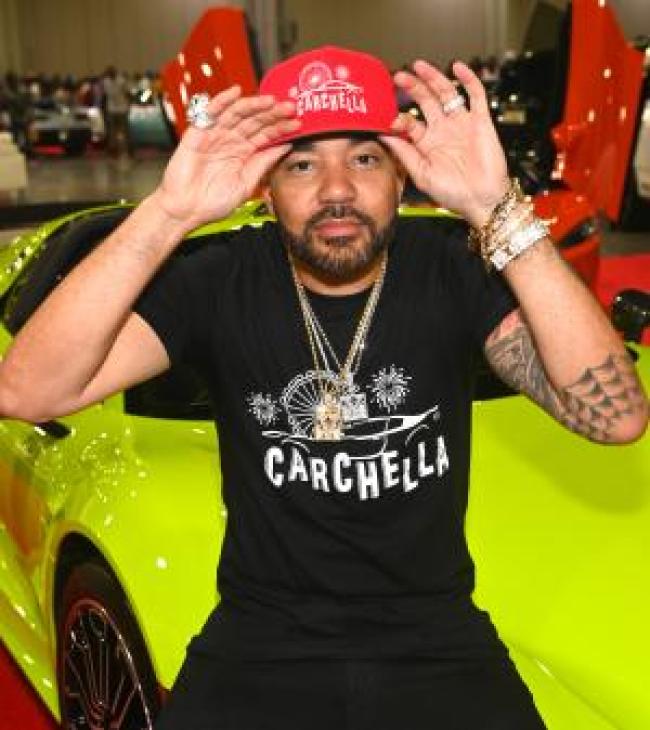 DJ Envy sosteniendo un sombrero rojo posando frente a un auto deportivo verde neon