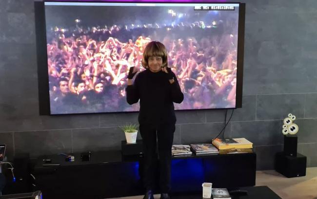Tina Turner de pie frente a un televisor en su casa con un traje negro.