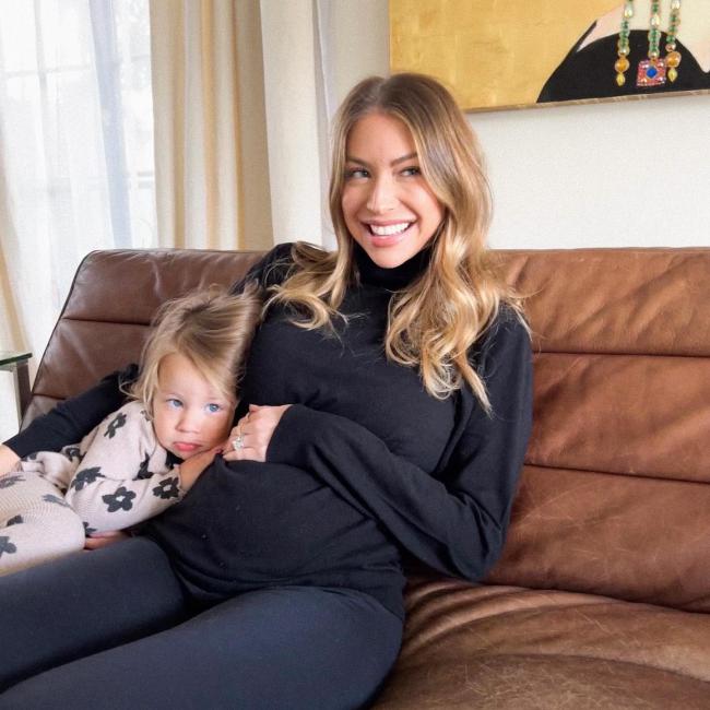 Stassi Schroeder embarazada acunando su barriga en un sofá con su hija Hartford.