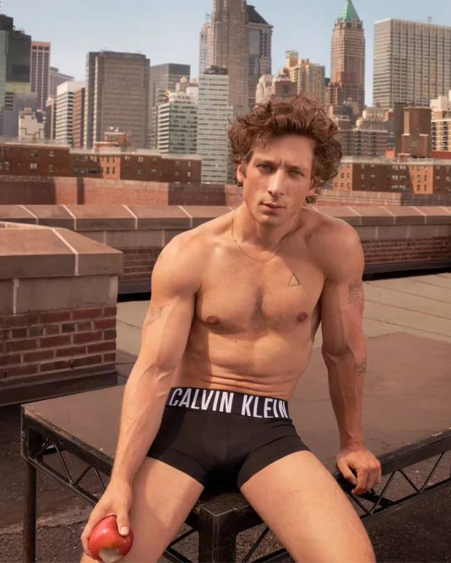 Jeremy Allen White en un anuncio de Calvin Klein, vistiendo solo calzoncillos, sentado afuera en una mesa y sosteniendo una manzana.