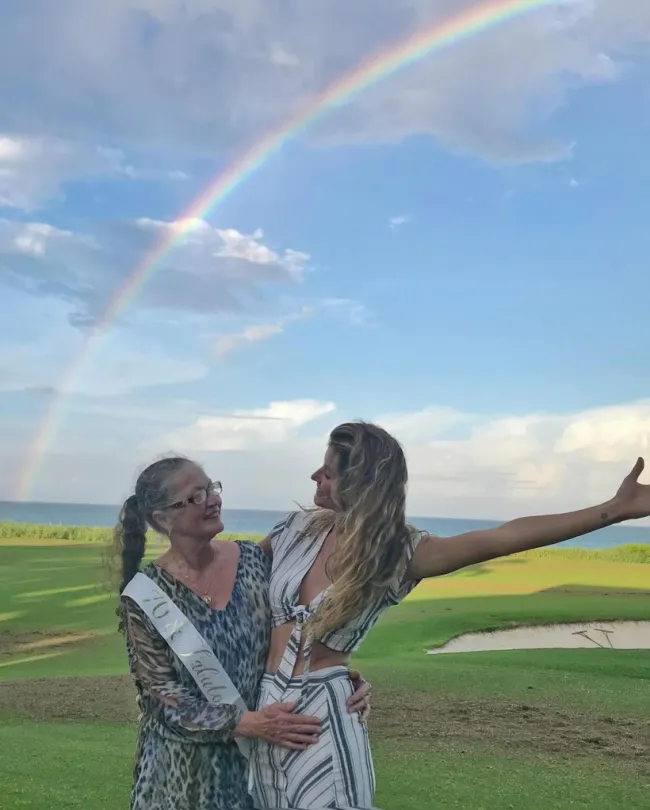 Gisele Bundchen y su mamá paradas en un campo de golf con un arco iris detrás de ellas.