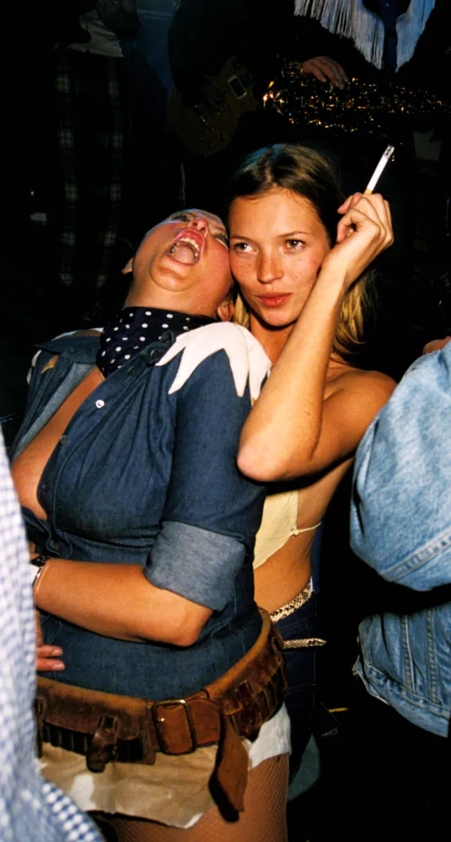 Kate Moss sosteniendo un cigarrillo mientras un amigo se inclina sobre ella.