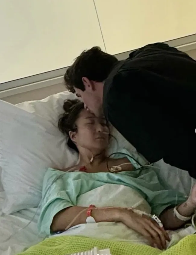 Ashley Park tumbada en una cama de hospital y su novio besándole la cabeza.