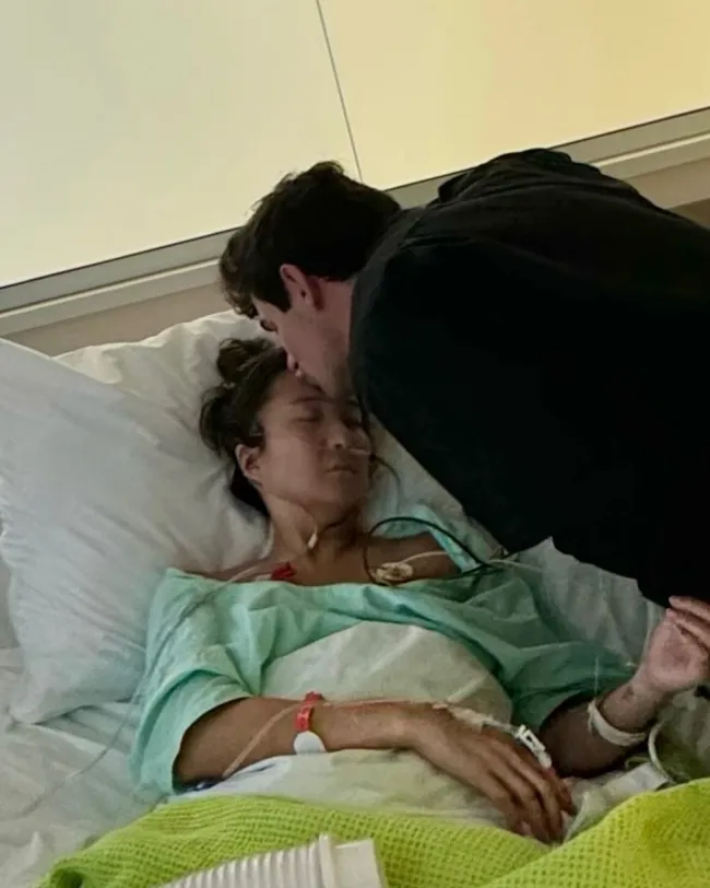 Ashley Park acostada en una cama de hospital y su novio besando su cabeza.