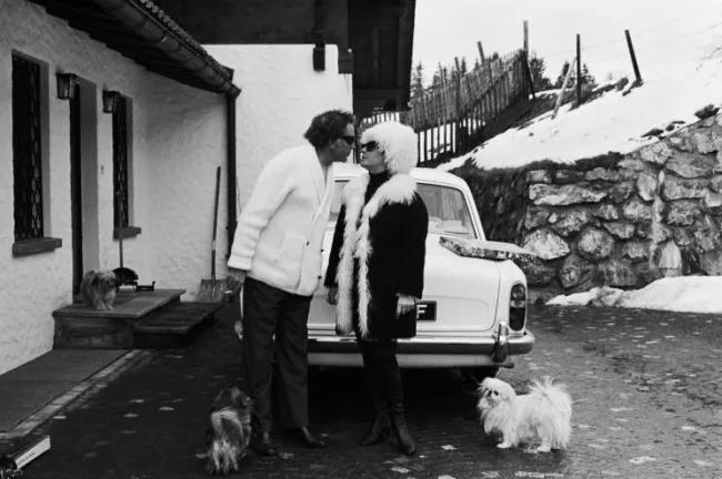 Richard Burton y Elizabeth Taylor besándose afuera de una casa y al lado de un auto. A su alrededor hay varios perros pequeños, con nieve al fondo.