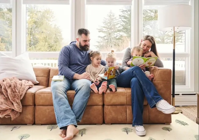 Jason y Kylie Kelce sentados en un sofá leyéndoles a sus tres hijas.