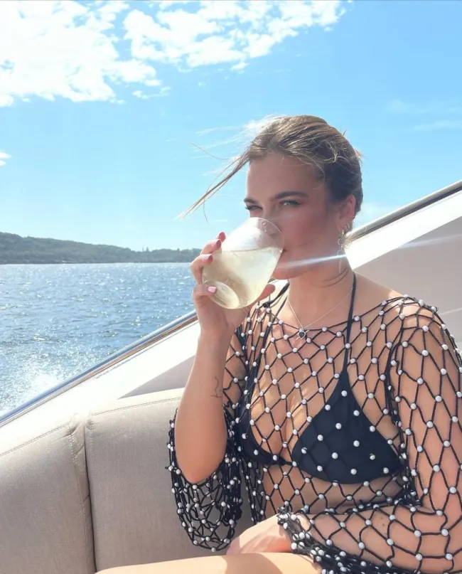 Stormi Bree con un bikini negro y una bata de crochet con perlas tomando una copa en un barco en Sydney.
