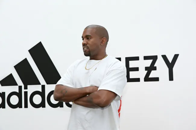 Kanye West de pie con los brazos cruzados frente a un cartel de Yeezy y Adidas.
