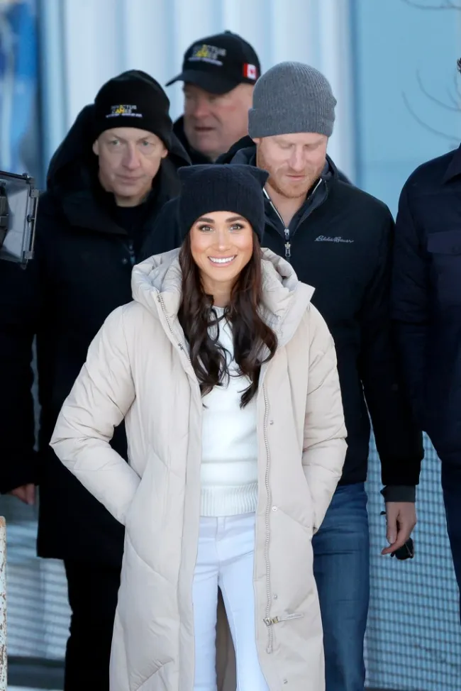 Meghan Markle sonriendo con sombrero, abrigo largo de invierno y jeans blancos. El príncipe Harry está detrás de ella.