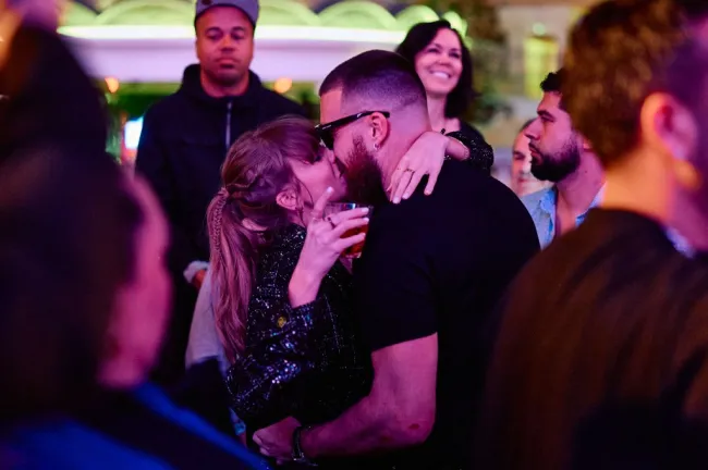 Taylor Swift y Travis Kelce se besan mientras ella sostiene una bebida.