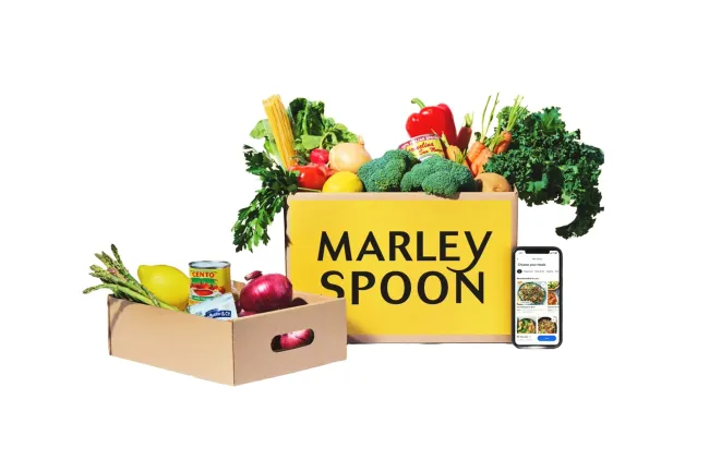 Una caja de Marley Spoon llena de productos agrícolas, con un teléfono abierto al sitio de la marca al lado.