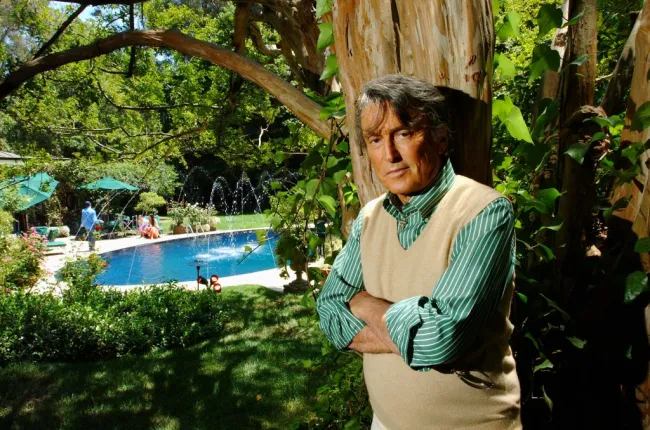 El productor Robert Evans apoyado contra un árbol frente a una piscina.