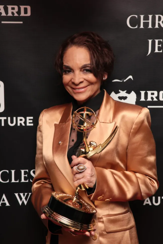 Jasmine Guy con camisa dorada sosteniendo el premio Emmy.