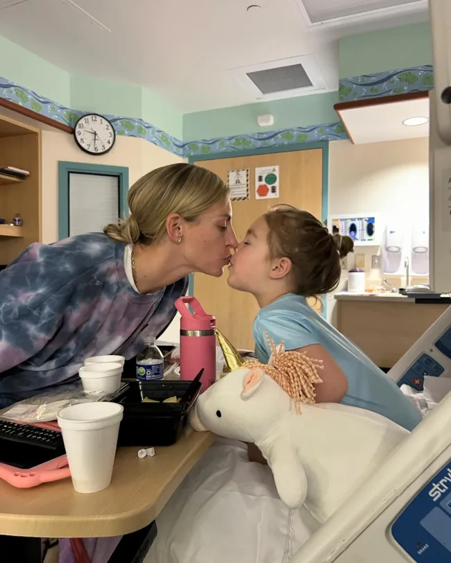 La esposa de Gunnar Peterson besa a su hija mientras ella se sienta en una cama de hospital.