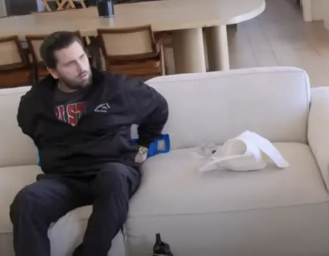 Scott Disick sentado en un sofá con una bolsa de hielo en la espalda.