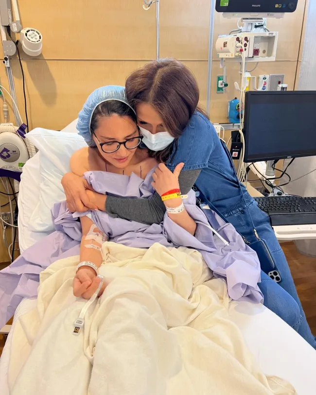 Olivia Munn en una cama de hospital con su mamá abrazándola.