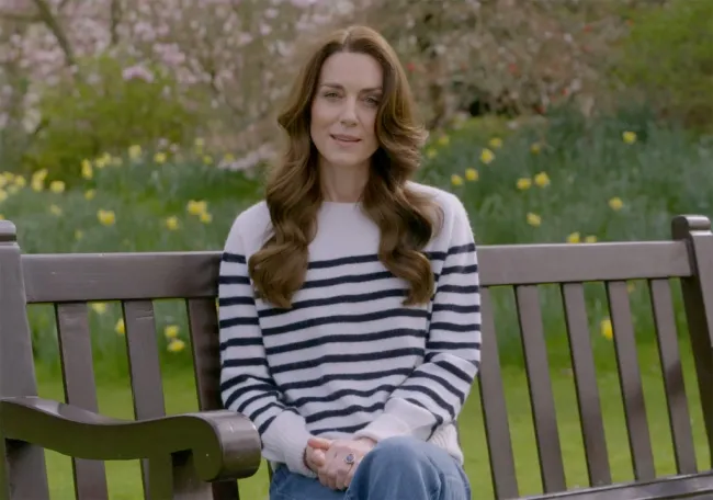 Kate Middleton sentada en un banco frente a flores y césped con un jersey de rayas