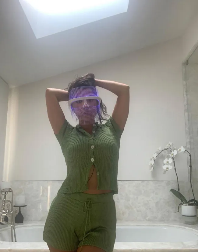 Lily Collins mirándose en un espejo mientras usa la máscara LED Current Body