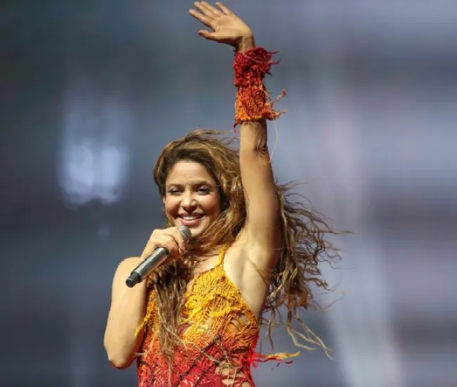Shakira con un top rojo y amarillo saluda mientras sostiene un micrófono.