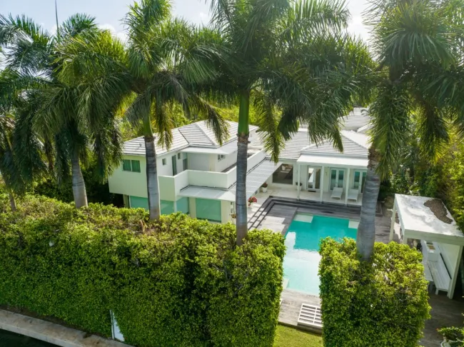Una casa tropical vista desde detrás de palmeras y setos. Tiene piscina y dos pisos en blanco.