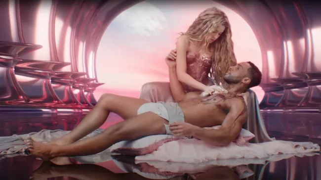 Shakira con un corpiño rosa inclinada sobre un hombre reclinado que parece tener sangre saliendo de una herida. Ella lo está frotando. Parece estar desnudo hasta quedar en ropa interior.