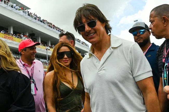 Tom Cruise con una polo blanca y aviadores de pie junto a Shakira, que lleva gafas de sol y una camiseta verde escotada. Están rodeados de hombres con gafas de sol y ropa informal y detrás se puede ver una tribuna.