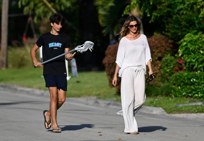 Gisele Bündchen vestida de blanco camina junto a su hijo que lleva un palo de lacrosse.